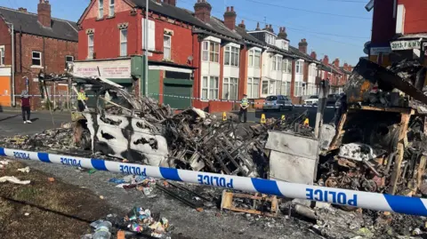 The charred remains of a burnt out bus on a public road, sat behind some police tape. Some terraced houses and police officers are stood in the background.