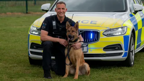 Hertfordshire Constabulary Sgt Dan Amos, holding on to PD Muzza, kneeling down in front of a yellow and blue marked police car, parked on green grass. Dan Amos is wearing all black and has short hair. 