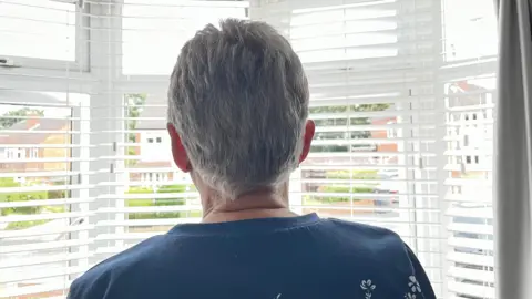 Woman seen from her back. She has short grey hair and is staring out of a window with blinds partially open. The view is of a well-maintained residential road. 