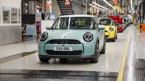 A line of differently coloured electric Mini cars are lined up on a production line in a factory. At least 10 cars can be seen inside the brightly lit and spotlessly clean factory.