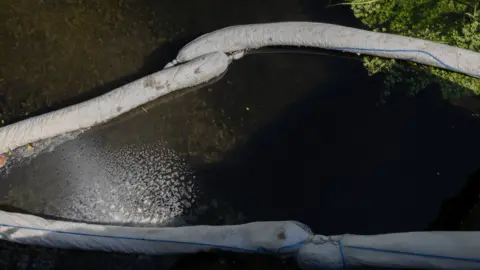 Facundo Arrizabalaga Long white sock-like containment booms hold paler water to the rest of the river.