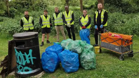 Peterborough Litter Wombles Six volunteers wearing yellow high-visibility jackets standing in a semi-circle holding their litter pickers. They are standing in front of plastic bags, with rubbish placed next to a black bin.