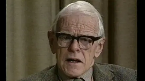 Éanna Mac Cana/Little Ease Films A still of Dr Denis Burkitt with his mouth open as he appears on a talk show. He is an older man, with white, medium length hair and is wearing a pair of black-framed square glasses. He is also wearing a light grey tweed suit jacket, with a light coloured collared shirt and a tie. Behind him is an olive green curtain.
