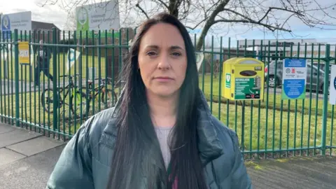 BBC/Seb Cheer Woman with long dark-coloured hair looks at the camera, with a neutral expression on her face. A school fence is in the background.