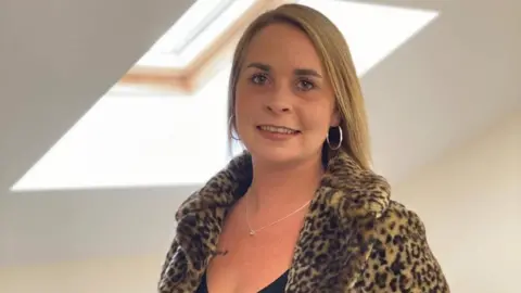 Charlotte Herring wearing hoop earrings and a necklace, as well as a furry leopard-print coat. She is smiling.