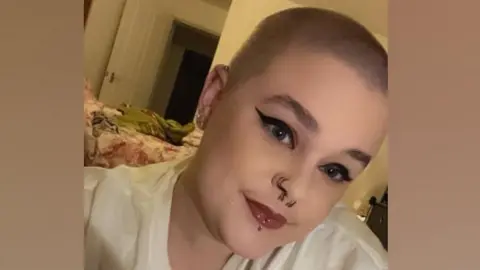 A head and shoulder picture of Tamzin Usher. She has a shaved head and blue eyes. She is wearing a white top and has piercings on her nose and lip. She has black winged eyeliner and dark lipstick. 