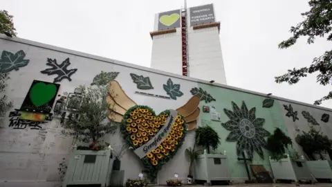 PA Media Grenfell tower covered in fabric and scaffolding, with a sign at the top that has a green heart and the phrase "Grenfell Forever in our hearts" on it. In the foreground are tribute to the victims hung and written on a nearby wall.