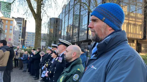 Men and woman in service uniforms and plain clothes stand in a line in Guildhall Square, looking to the left.