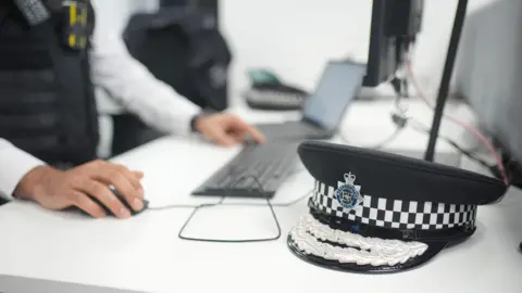 Police hat in foreground next an officer using to keyboard, laptop and computer screen in background