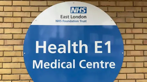 The front of a homeless medical centre run by East London NHS Foundation Trust in London. The picture shows a sign that says Health E1 Medical Centre with a brick wall behind it