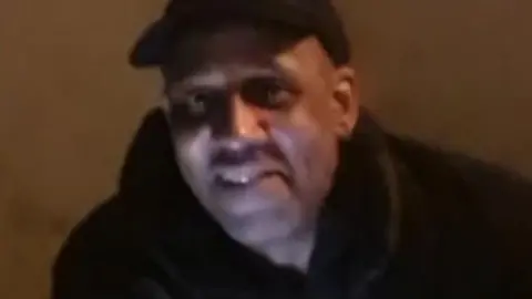 A man in low lighting smiles. He is wearing a black top and a black cap.