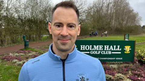 A man with short dark hair, wearing a tight light blue zip up golf jumper, is pictured by a sign for Holme Hall Gold Club. Also in the background are several golfers, trees and shrubbery.