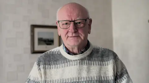 A balding man looks at the camera. It is a head and shoulders shot. He has black-rimmed glasses and is expressionless. He has a grey and white jumper on. Behind him are white walls with a framed photo on the wall.