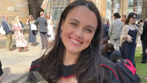 Alex Easson Alex wearing a graduation gown with red trim stands outdoors in a courtyard surrounded by historic stone buildings and large windows, with groups of people in formal clothing gathered and talking in the background.
