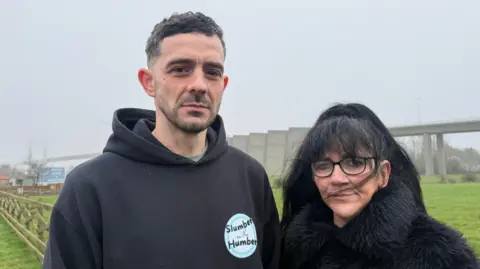 A man and a woman stand in front of a suspension bridge on a windy, foggy day. The man, on the left, is wearing a black hoodie. He has brown hair and a brown beard. The woman, on the right, is shorter and wearing a black fluffy coat. She has black hair and glasses. Only parts of the bridge are visible in the thick fog. 