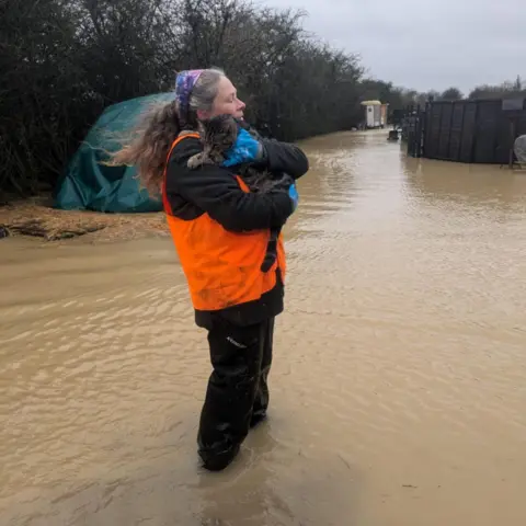 Fairfield Community Farm Kathy Owen wearing an orange hig-vis vest standing in flood water and holding a cat