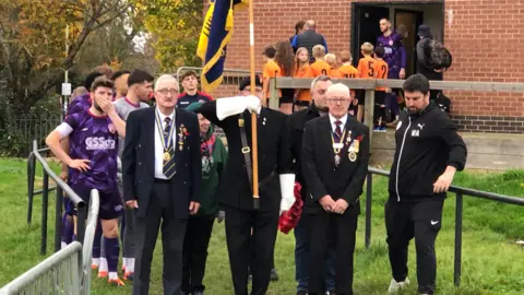 Three veterans carrying a flag lead out the teams at a football match