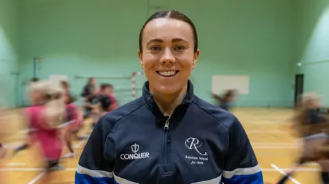 A woman smiling at the camera. She is wearing a jumper which says "Rainham School for Girls" on it. There are children running behind her.