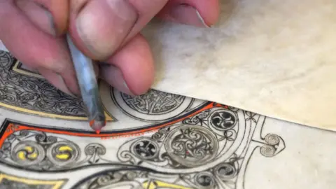 Norman Strachan A close up of Thomas Keyes' fingers holding a drawing tool while colouring in motifs on vellum 