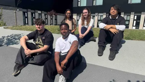 BBC Five students sit on a hill outside Cambourne Sixth Form College. There are two girls and a boy on the back row and two boys on the front row. They are all looking at the camera. It is a sunny day and they cast shadows onto the ground around them. 