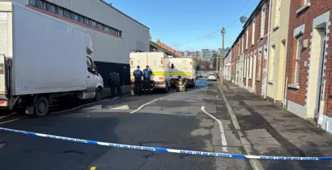 Police are at the scene of a security alert on Empire Street, south Belfast where residents have been evacuated. The sky is blue and there is a cordon. There is a red brick wall and a police cordon and police vans. 