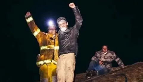 Two men stand on a roof with arms raised smiling at the camera. One is wearing a high vis suit and the other a dark jacket stained with ash. Another man sits behind them wearing a lumberjack shirt