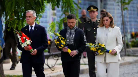 The Canadian Press via AP Canada's Prime Minister Mark Carney, his grey hair short and face sombre, holds red and white flowers as he walks in a dark suit on a street in Kyiv, Ukraine. Ukrainian President Volodymyr Zelensky, his hair and beard dark and short, wears a black traditional Ukrainian outfit with white embroidery around the collar and in a line down the front. His wife, Olena Zelenska, wears a white suit. The couple wear sombre expressions and both carry yellow and blue flowers. A soldier walks in the background.