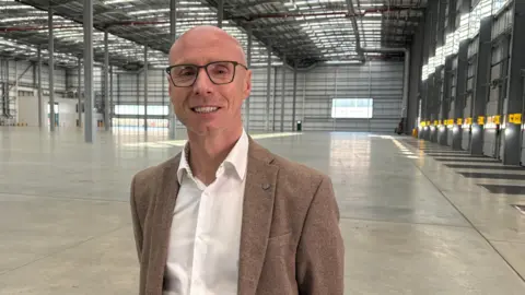 Ben Schofield/BBC Dan Myers looking directly down the camera standing inside a large empty warehouse. He is wearing a white, open-necked shirt and a light brown coloured blazer. He also has black, rectangular framed glasses on. He is smiling, showing his teeth, and is bald. 