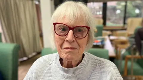 Shelagh has short white hair and red rimmed glasses.