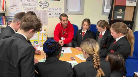 The image shows a group of school pupils in uniform gathering around a table in a classroom while an adult in a red hoodie sits with them, looking at worksheets and learning materials.