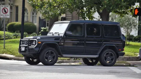 A a black Mercedes Benz G Wagon on a road ear a tree, grass verge and buildings in the background