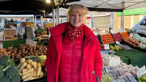 Woman wearing a bright pink coat and scarf smiling at the camera, stood in front of a fruit and veg stand