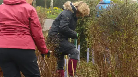 Rodborough Parish Council A woman is digging a hole with a shovel between bushes. She is wearing a rain coat with a furry hood and wellies.