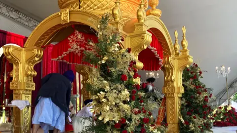 A ceremonial display of the Guru Granth Sahib, the Sikh holy scripture, placed under an ornate golden canopy. The setting resembles a temporary darbar (prayer hall) created for Vaisakhi celebrations. Rich red drapery and elaborate floral arrangements frame the space. Two individuals in turbans kneel in reverence, reflecting the deep respect shown to the scripture during Sikh festivals.