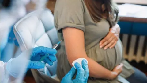 Getty Images A pregnant woman in a green dress, holding her bump, sat in a grey chair, while the hands of a medical professional in blue gloves prepares a vaccine. 