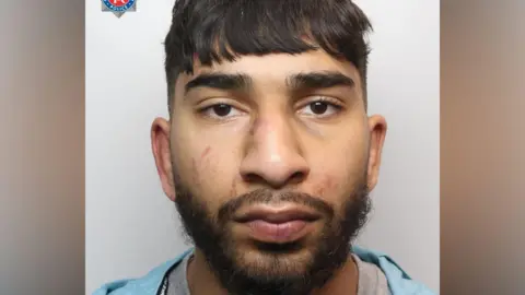 A mugshot of an Asian man in his early 20s, who has black hair cut into a fringe and black facial hair. He has red marks to his face and is looking into the camera with a blank expression. 