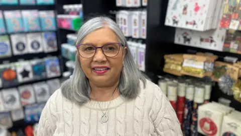 Photograph of Meena Sheth, who runs a card store in Bramhall. She is pictured inside her shop. With long grey hair and wearing purple glasses, she is wearing a cable-knit woollen jumper.
