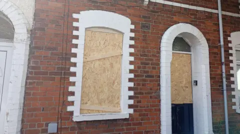 A red brick house, the door and front window are boarded up.
