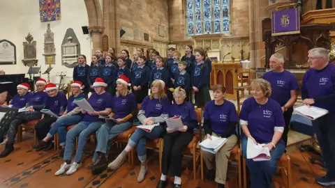 The choir singing. There are school children standing behind the adults. The adults are all wearing purple t-shirts and some are wearing Santa hats.