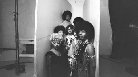 Getty Images A black and white image of Sly and the Family Stone. They stand between two white boards, close to one another.