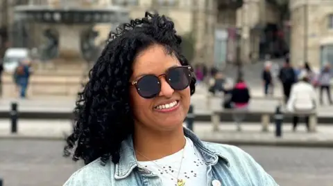 Michela stands in piazza smiling wearing dark glasses. Her black curly hair skims  her shoulders and is blown over to the side by the wind.
