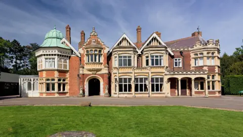 PA Media The Bletchley Park manor house. It is a two-storey building with large windows and red bricks. A grass lawn is in front of the manor along with a a driveway.