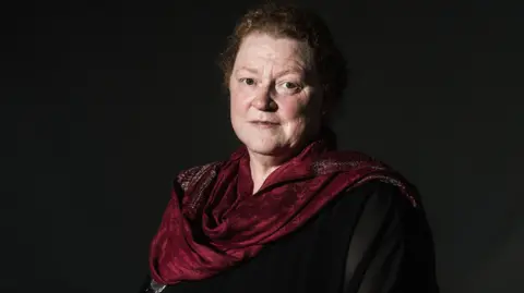 Getty Images A lady with ginger hair stares at the camera with a serious expression. She is wearing a dark blouse and a dark red scarf.