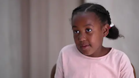 Eden is wearing a pink top. Her hair is plaited in two braids, held at the end with a white bobble hair band.