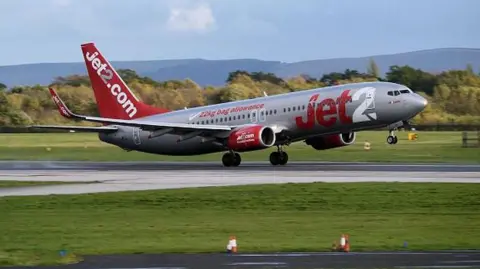 Jet2 aircraft takes off from a runway at Manchester Airport on a clear day. Hills of the Derbyshire Pennines in the background.