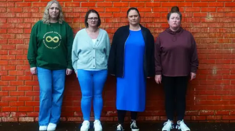 BBC Four women. Far left has blonde hair and is wearing a green sweater and blue jeans, next to her has shot brown hair and is wearing a sky blue cardigan and blue jeans, next to her has brown hair pulled back into a pony tail, a blue dress and darker blue jacket and next to her has brown hair in a top knot, a maroon hoody and dark blue leggings
