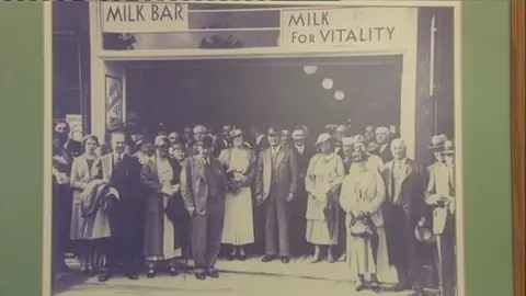 A crowd of well dressed people stand outside a milk bar 