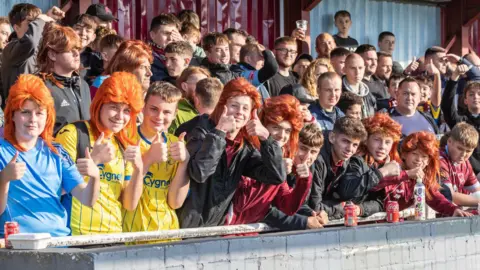 Taunton Town Football fans wearing orange mullet wigs. They are in the stands and wearing football merch.