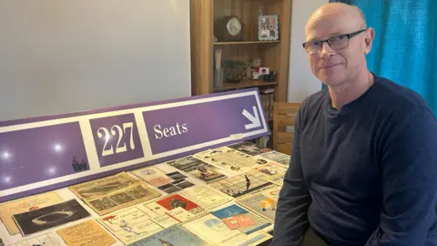 Graham Cooksley sat in his kitchen in front of a selection of his collection which is displayed out on the table. They are a lots of programmes, tickets and books as well as a big purple original Wembley stadium seat sign. 