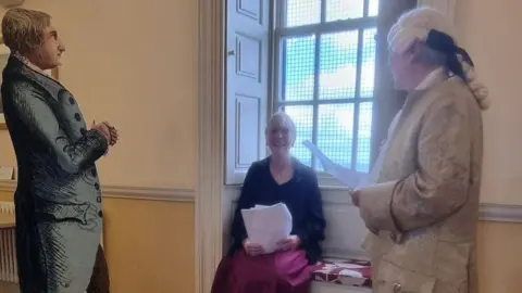 Rotherham District Civic Society A historical indoor scene shows two costumed figures in period clothing, with one reading from a sheet of paper near a large window. A seated person holding papers is positioned between the figures, while a life-size illustrated cutout stands on the left side of the room. The setting features cream-colored walls, natural light from the window, and furnishings that suggest a heritage or museum environment.